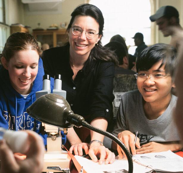 Diverse group of students seated at a table with a female instructor smiling as they lean between the students.
